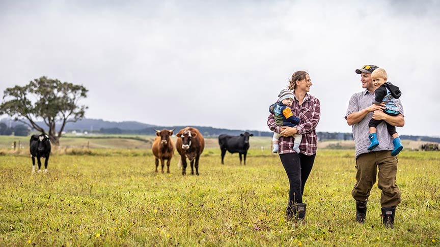 farming couple both holding young children walking in paddock with cows 