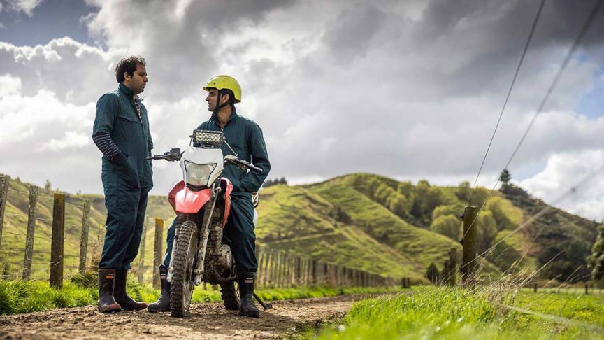 Man with hands in pockets of jumpsuit talks to man sitting on dirt bike 